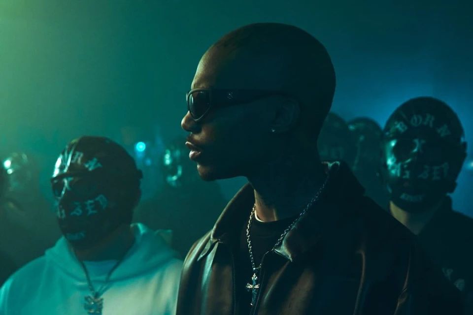 Close up of an adult male wearing sunglasses and a chain, surrounded by a crowd with masks, against a blue-green background.