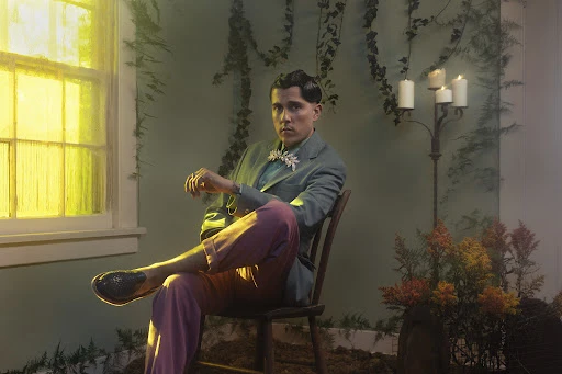 Well-dressed man poses indoors on wooden chair wearing a teal jacket and pink pants, with a bow tie, set against a plant-covered wall.