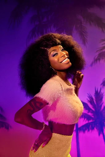 A smiling African-American woman with an afro hairstyle poses in a studio wearing a fuzzy crop top and skirt against a tropical backdrop.