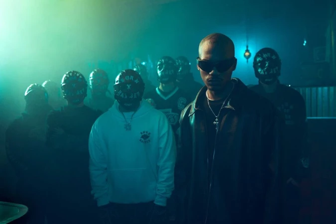 Music video-style portrait of a bald adult male in sunglasses and leather jacket, with a group of masked people in the background.