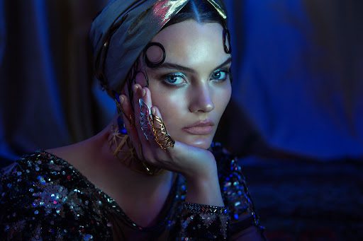 Close-up portrait of a woman with dramatic makeup, shimmering head wrap, and ornate rings, set against a draped, dark blue background.