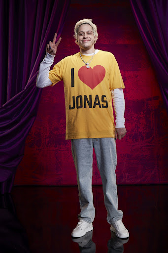 Full shot of adult man wearing a yellow t-shirt with the words 'I Heart Jonas', blue jeans, and white sneakers in a studio setting.
