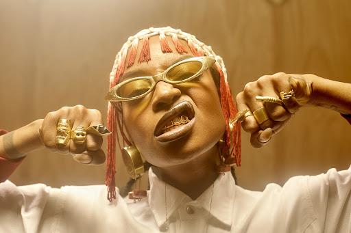 Portrait of a woman with gold grillz, wearing gold sunglasses and ornate gold rings, indoors in a photo studio with warm lighting.