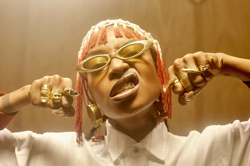 Portrait of a woman with gold grillz, wearing gold sunglasses and ornate gold rings, indoors in a photo studio with warm lighting.