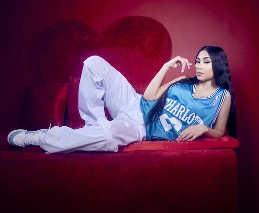 Young woman in a teal basketball jersey and white pants poses against a red backdrop with a heart-shaped pillow.