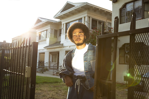 Adult male with afro stands in front of white clapboard houses and a black iron fence on a sunny day, wearing a denim jacket.