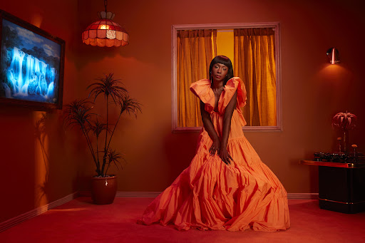 A woman in a ruffled orange dress poses in an orange room with retro decor, including a glowing lamp and a landscape painting.