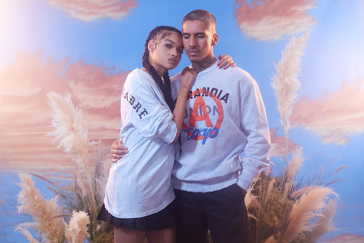 A stylish couple poses in front of a sky and feather background, both wearing casual sweatshirts and black bottoms in soft light.