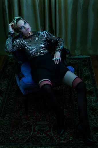 A woman poses while seated on a chair, wearing a sheer star-patterned top, dark skirt, black thigh-highs, and jewelry in an indoor setting.