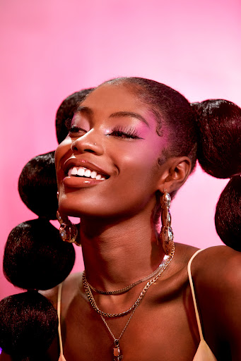 Smiling African-American woman poses against a pink backdrop showcasing a unique braided hairstyle, bold makeup, and gold jewelry.