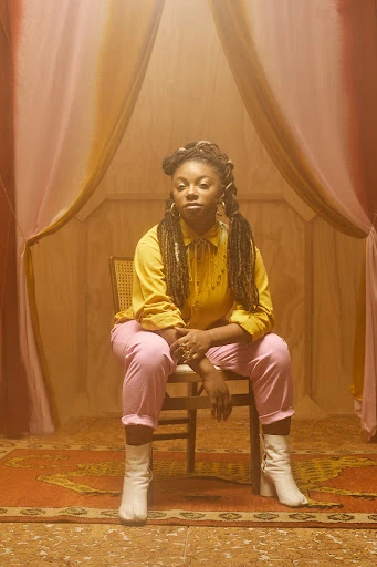Stylish woman with braids sitting on chair, wearing a yellow top and pink pants in a warm-toned, tented photo studio.
