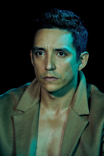 Portrait of a person with dark hair wearing a tan coat, set against a dark backdrop with teal and blue lighting accents.