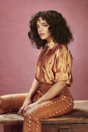 Portrait of Tessa Thompson wearing a glossy copper satin shirt and matching trousers seated on a wooden box