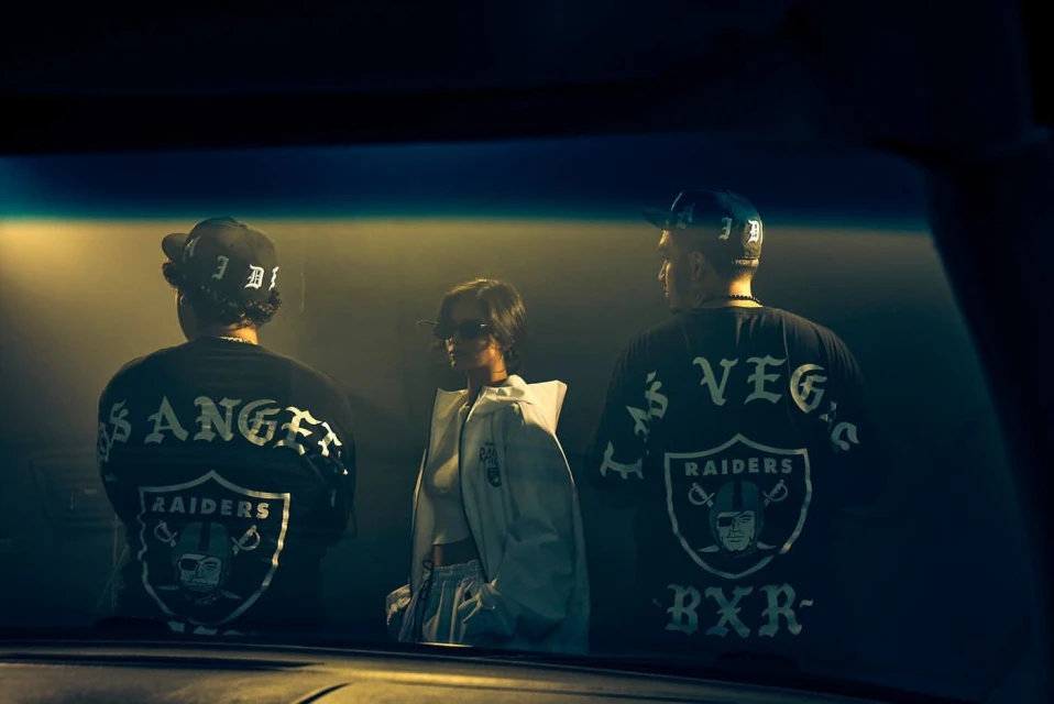 Three people wearing Los Angeles Raiders and Las Vegas Raiders shirts, standing in front of a vehicle windshield at night.