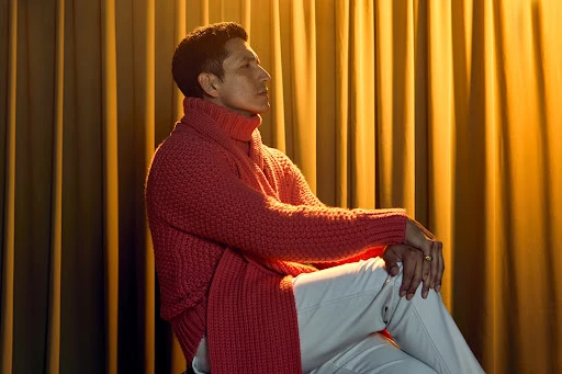 Man in a red turtleneck sweater and white trousers seated against warm gold draped curtains in a portrait by Gizelle Hernandez.