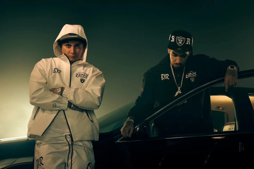 Two adult males pose next to a luxury car while wearing matching 'Raiders' themed apparel. The setting is a dark studio environment.