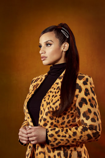 Portrait of a woman with brown hair wearing a leopard print blazer against a brown background, looking off to the side.
