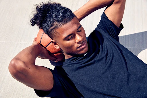 Athletic man does crunches with a basketball behind his head, resting on a light grey surface in natural light.