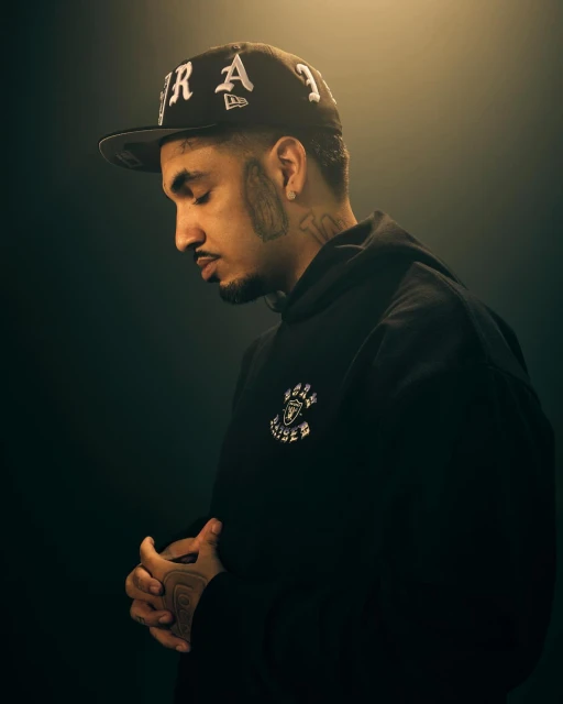 Portrait of a man with a baseball cap and hoodie in a dark studio, hands clasped, soft lighting on the face and upper body.