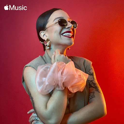 Portrait of smiling adult female model with sunglasses, stylish jewelry, and a large hair scrunchie against a vivid red background.