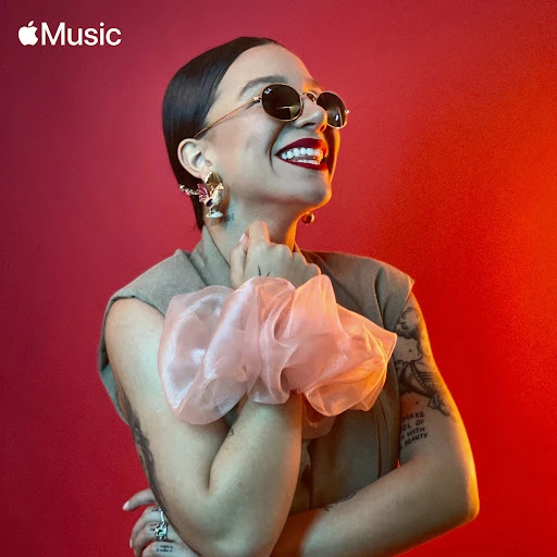 Portrait of smiling adult female model with sunglasses, stylish jewelry, and a large hair scrunchie against a vivid red background.