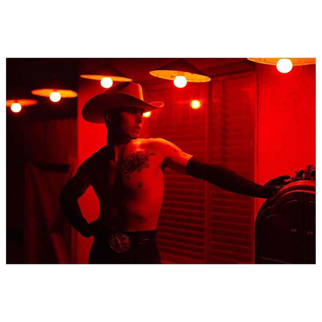 Shirtless man in cowboy hat poses indoors with red lighting. He wears black pants and gloves, with arm tattoos visible.