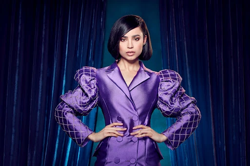 Sofia Carson posing in a purple blazer with ornate puffed sleeves against a dark blue curtain backdrop in a studio setting.