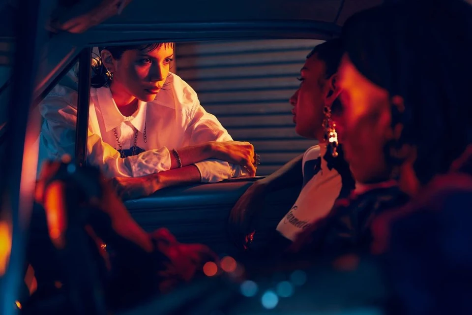Three women in vintage fashion inside a car at night, illuminated by vibrant orange and blue lighting, evoking a retro aesthetic.