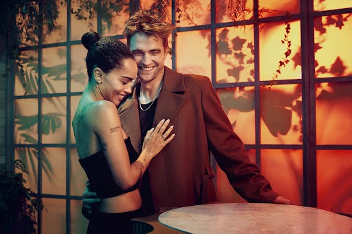 Smiling couple embraces in a warm-toned photo studio setting with orange and teal lighting, foliage, and a grid-patterned window.