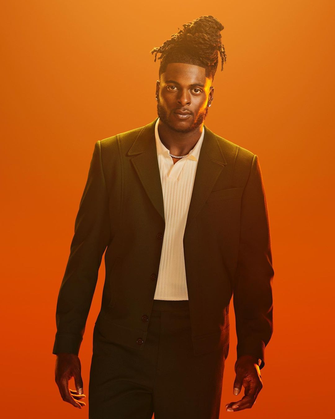 Portrait of a Black man wearing a dark suit and light-colored collared shirt against a bright orange background in a medium shot.