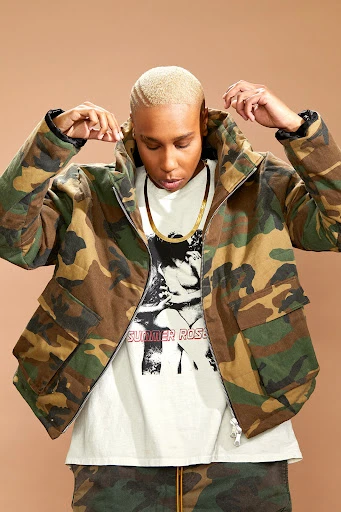 Adult in a camo jacket stands indoors against a neutral backdrop, wearing a white tee with a graphic and a gold chain necklace.