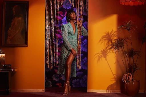 A Black woman poses in a turquoise coat dress against an orange wall, accentuated by a flower curtain, lamp, and a vintage painting.