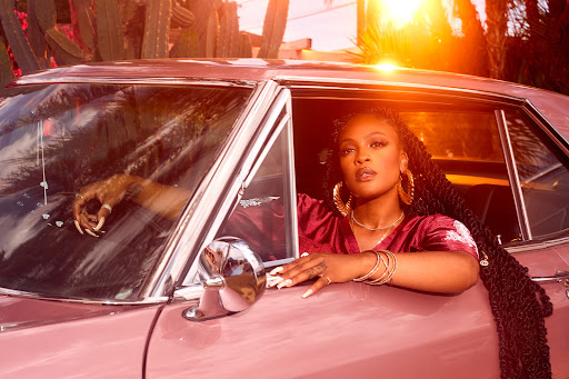 A woman with long braids and gold hoop earrings leans out the window of a pink classic car with cacti in the background.