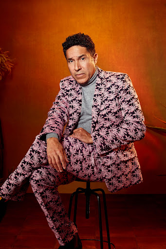 An adult male sits on a stool with a patterned suit, his expression neutral, set against an orange studio backdrop with plants visible.