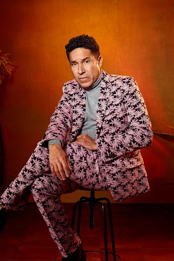 An adult male sits on a stool with a patterned suit, his expression neutral, set against an orange studio backdrop with plants visible.