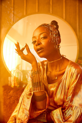 Elegant African-American woman in shimmering gold outfit and gold jewellery poses indoors in front of vintage furniture and decor.