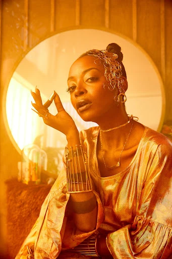 Elegant African-American woman in shimmering gold outfit and gold jewellery poses indoors in front of vintage furniture and decor.