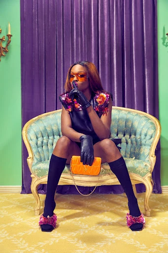 A fashionable Black woman sits on an ornate turquoise chair in a photo studio, wearing a black outfit with floral accents and orange sunglasses.