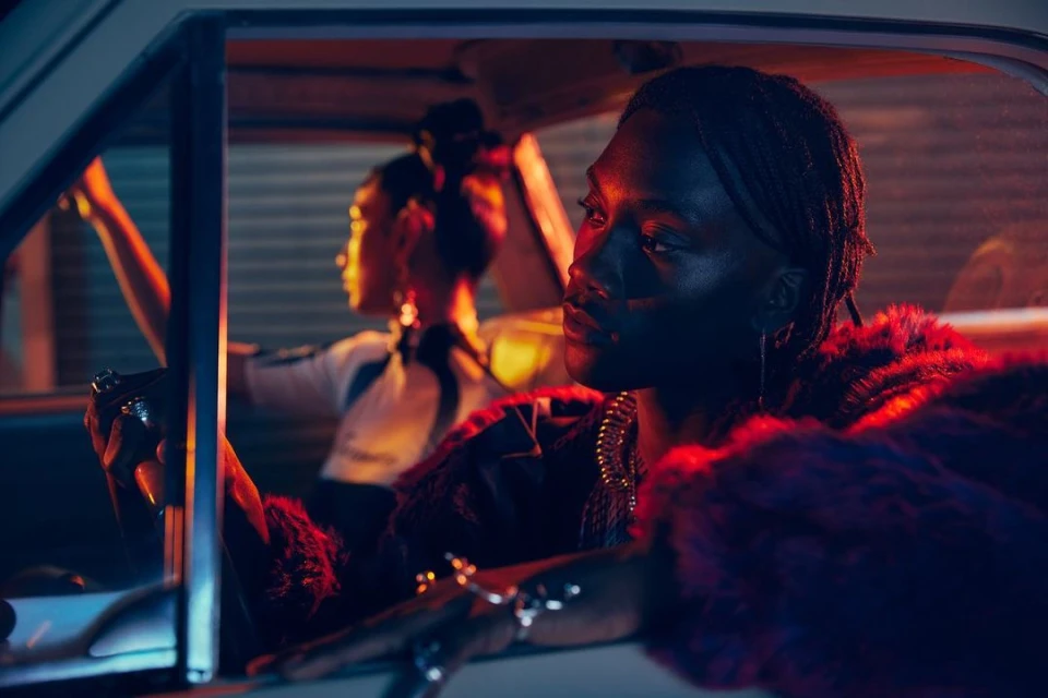 Two people in a car at night, focus on the driver wearing jewelry and a fur jacket, with another person visible in the back seat.