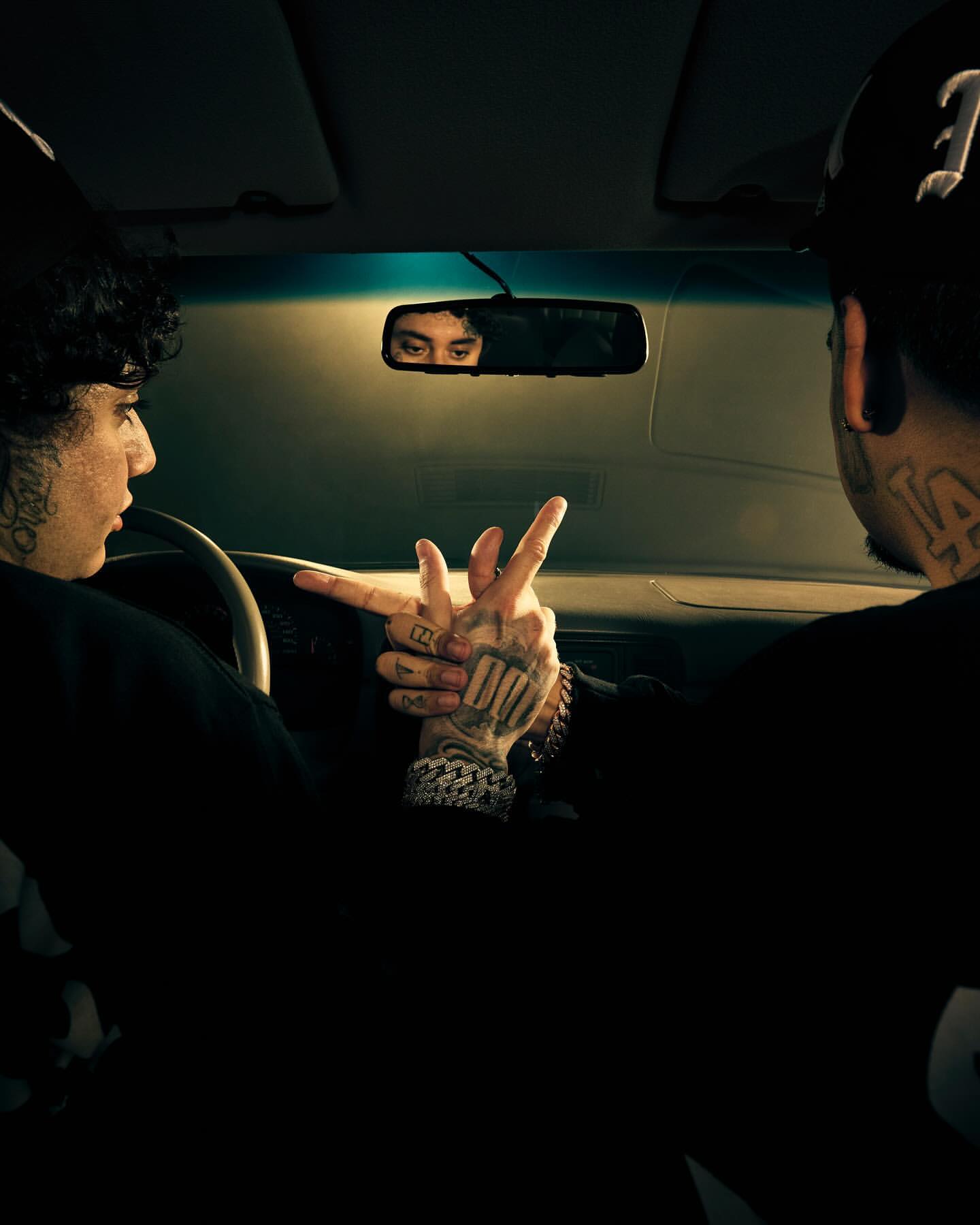 Two men inside car displaying tattooed hands with gestures in dark interior, focus on hand tattoos and mirrored eyes in rearview mirror.