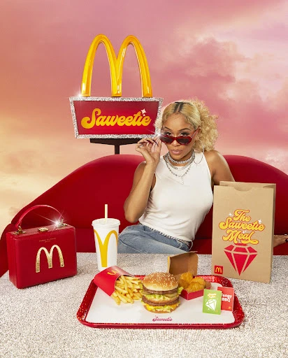 Promotional photo of Saweetie next to a McDonald's meal, with a large McDonald's sign and purse, set against a pink cloudy sky.