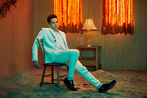 Man in a white suit seated on a wooden chair in a room with a sand floor and wood-paneled walls by Gizelle Hernandez