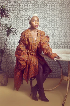 Cynthia Erivo wearing a plaid coat and tall boots seated at a table against a patterned wall, photographed by Gizelle Hernandez.