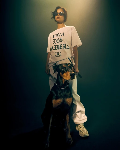 A person stands with their doberman pinscher in a studio, wearing a white t-shirt, sweatpants, sunglasses, and chunky sneakers.