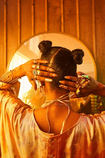 Back view of a woman with hair styled in space buns, accessorized with jewelry, in front of an oval mirror and wooden background.