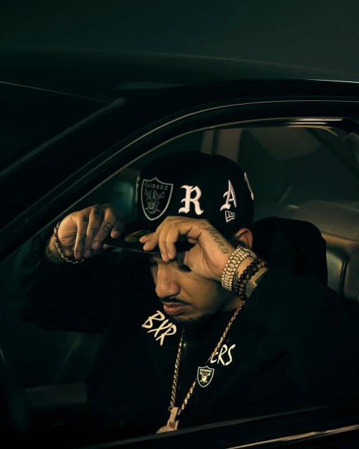 Adult male in driver's seat, wearing a black Raiders baseball cap and sweatshirt, gold jewelry, in a car with dark, moody lighting.