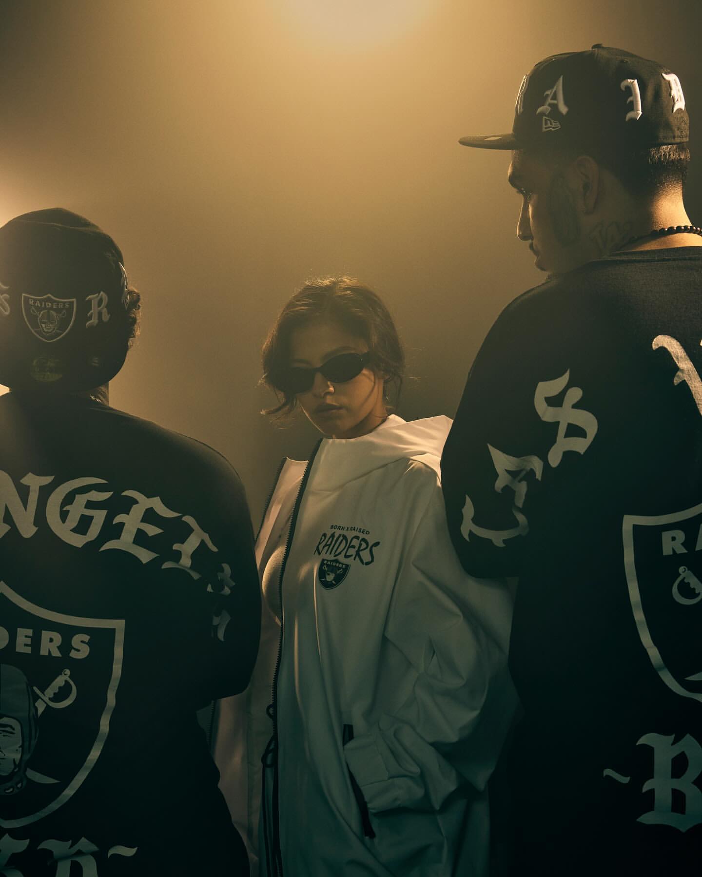 Three people in 'Los Angeles Raiders' fan apparel stand indoors. The woman in the middle wears sunglasses and a white jacket.