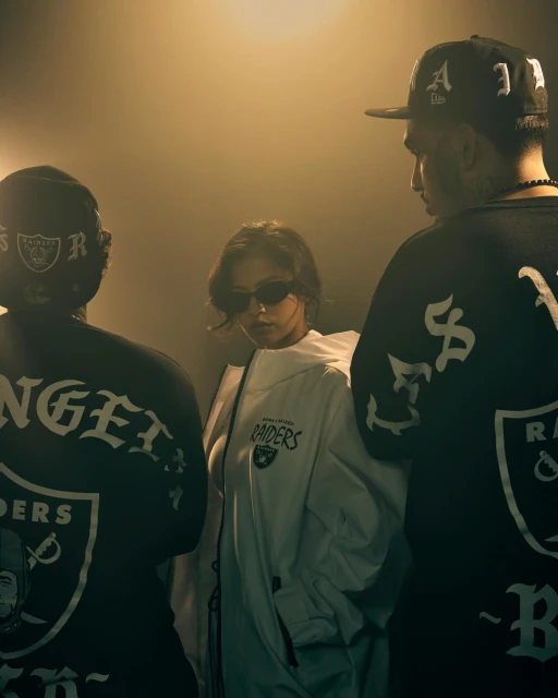 Three people in 'Los Angeles Raiders' fan apparel stand indoors. The woman in the middle wears sunglasses and a white jacket.