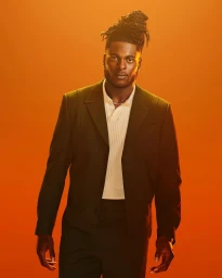 Portrait of a Black man with dreadlocks in a bun wearing a dark suit jacket and white ribbed polo shirt against a warm orange background.