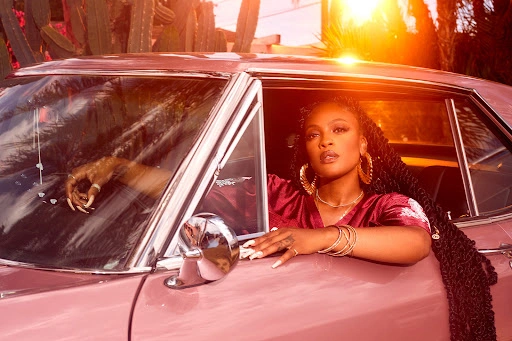 Black woman with braided hair wearing a pink top leaning out of the window of a vintage pink car during sunset